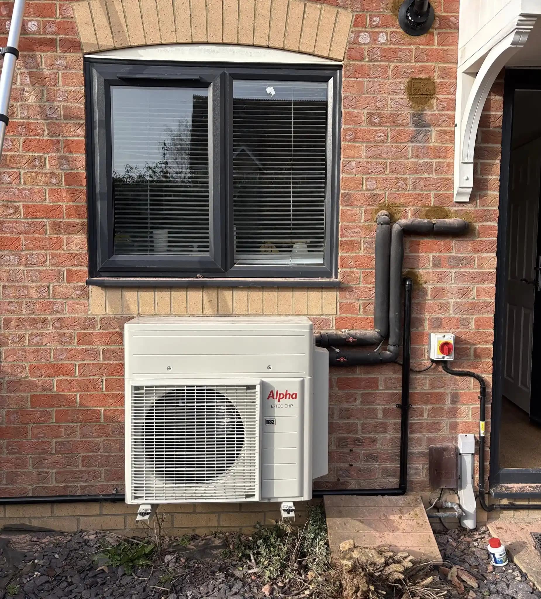 Alpha heat pump outdoor unit installed on an exterior brick wall.